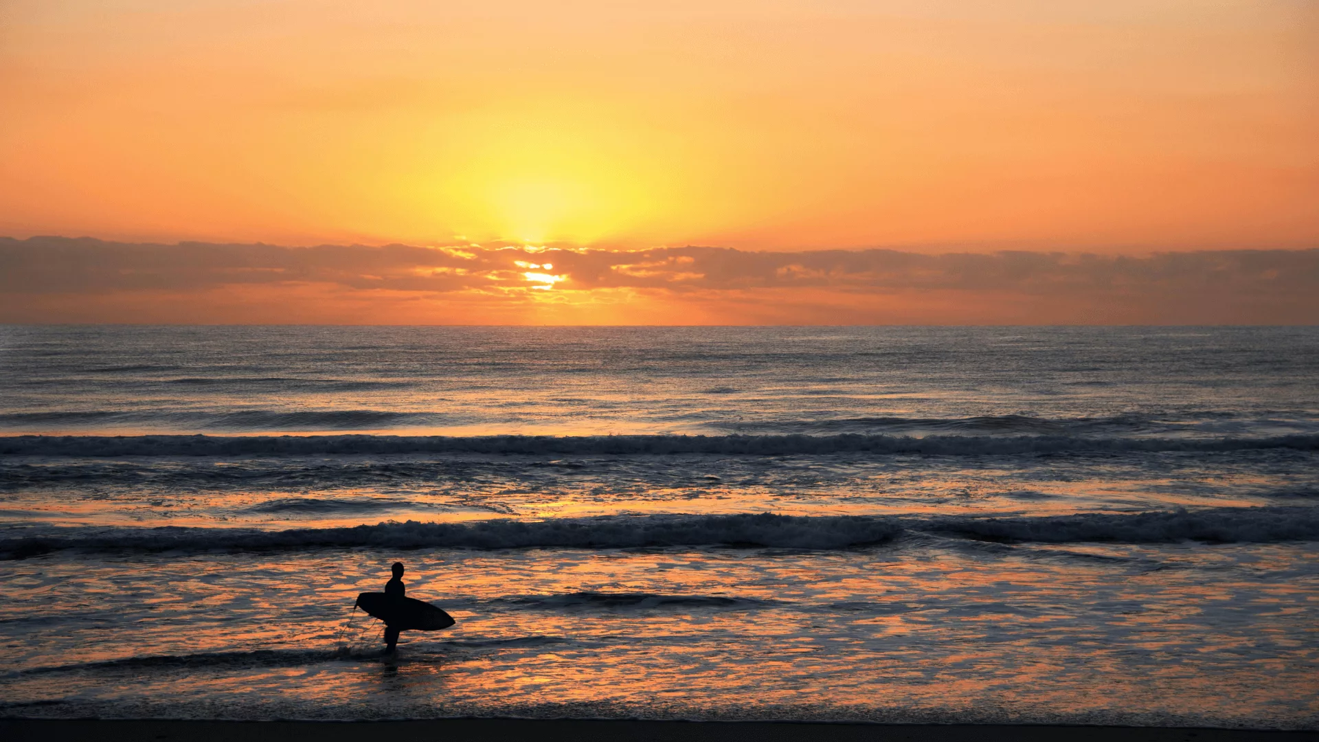 A photo of a warm sunset over the ocean. In the bottom left hand corner is a surfer holding a surfboard looing out at the horizon