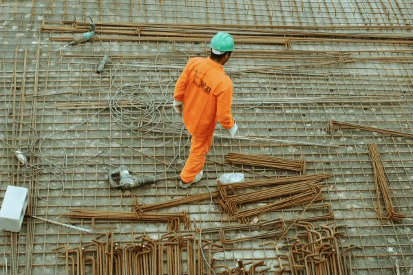 construction worker wearing a bright orange jumpsuit and a green hard hat, walking across a metal grid foundation, which is covered in rebar and construction tools.